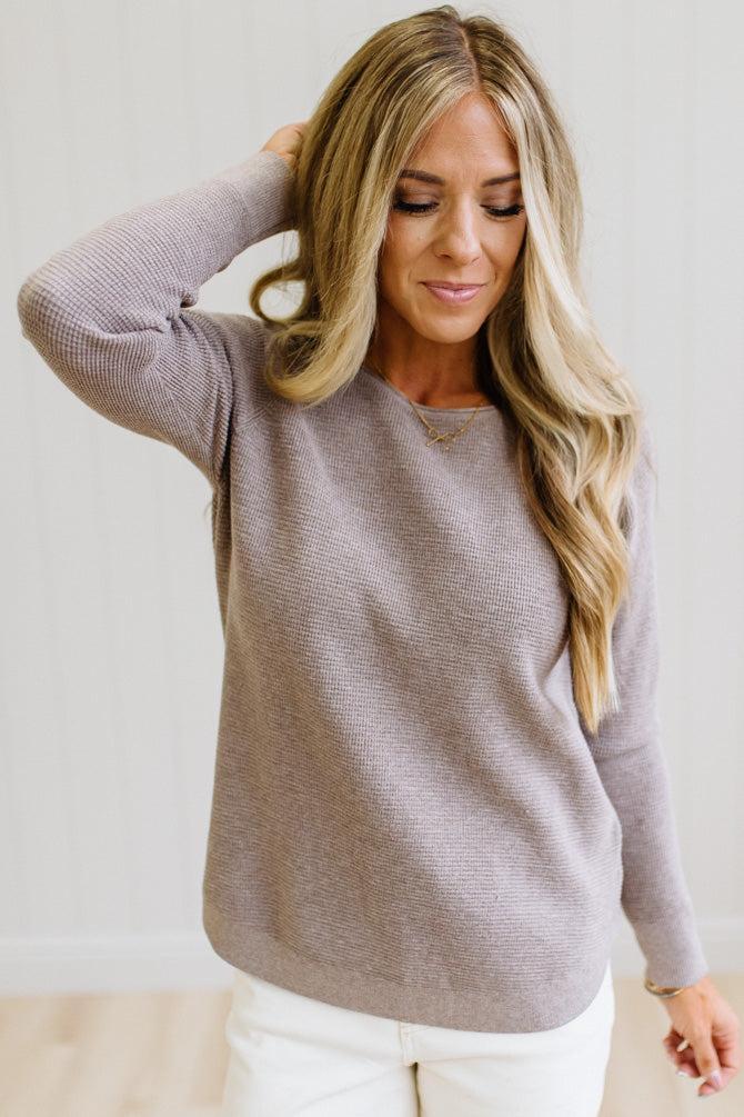A woman wearing a ribbed knit long-sleeve sweater in a muted taupe-gray color, paired with white pants, standing against a light neutral wall.