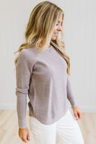 A woman wearing a ribbed knit long-sleeve sweater in a muted taupe-gray color, paired with white pants, standing against a light neutral wall.