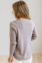 A woman with blonde hair is shown from behind wearing a light taupe ribbed knit sweater and white pants, standing against a white paneled wall.