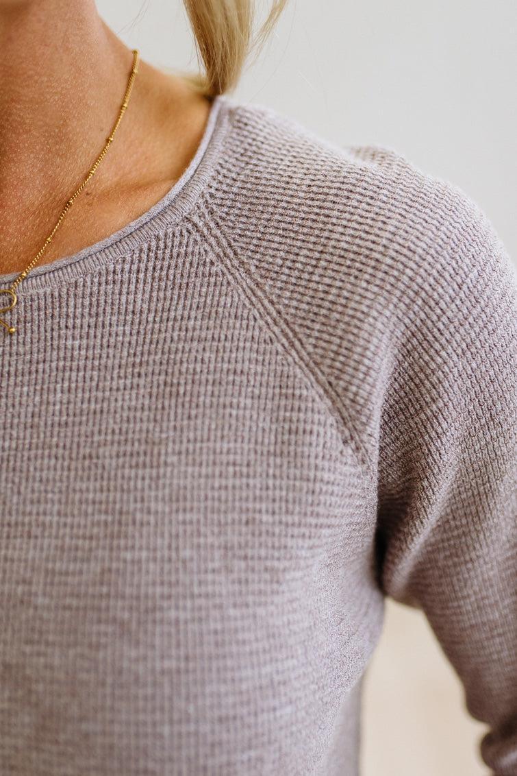 Close-up of a person wearing a ribbed knit crewneck sweater in a light taupe-gray color, paired with a delicate gold chain necklace.
