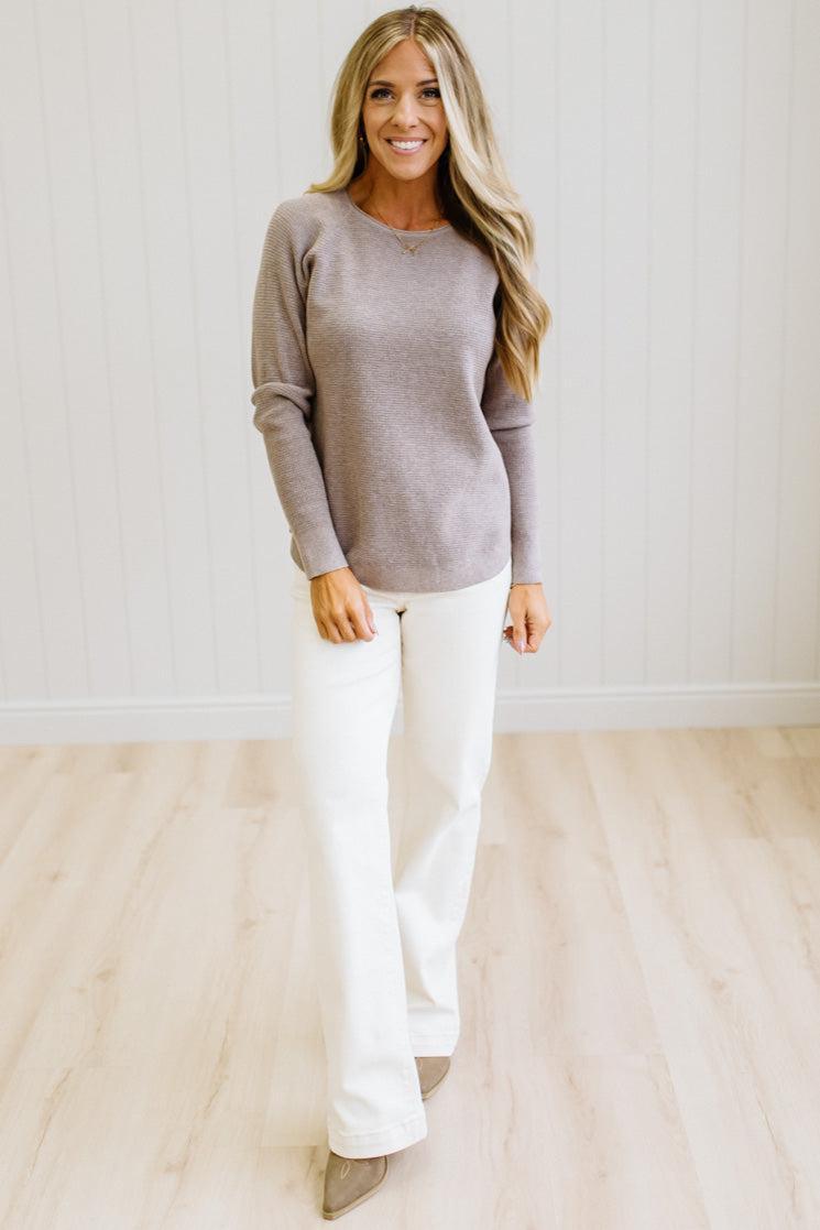 A woman modeling a light taupe long-sleeve knit sweater and white wide-leg pants in a bright room with white paneled walls and light wood floors.