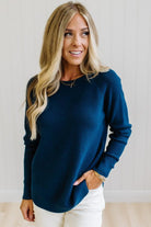 A woman with blonde hair wearing a dark navy blue ribbed knit long-sleeve top and white pants, standing against a white paneled wall.