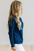 A woman with blonde hair wearing a dark navy blue long-sleeve knit sweater and white pants, standing against a white paneled wall.