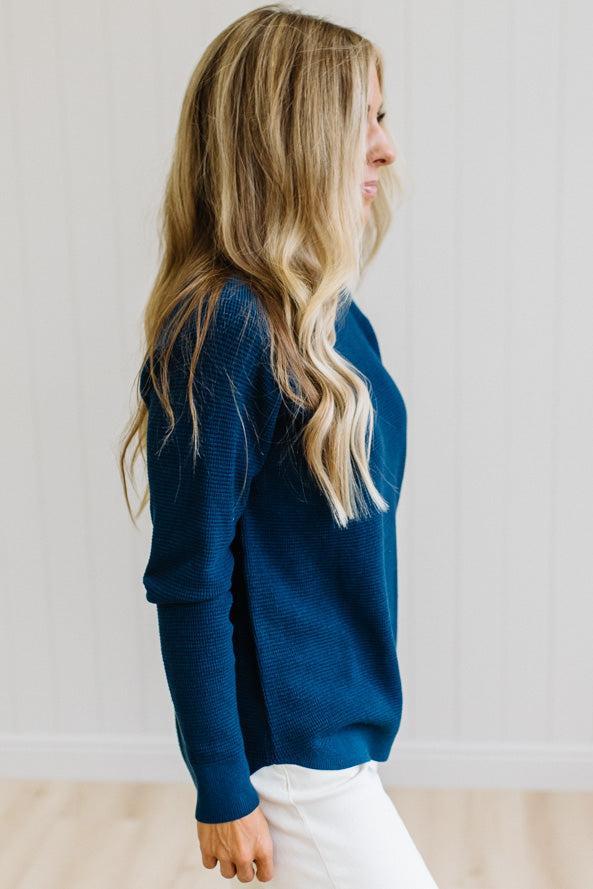 A woman with blonde hair wearing a dark navy blue long-sleeve knit sweater and white pants, standing against a white paneled wall.
