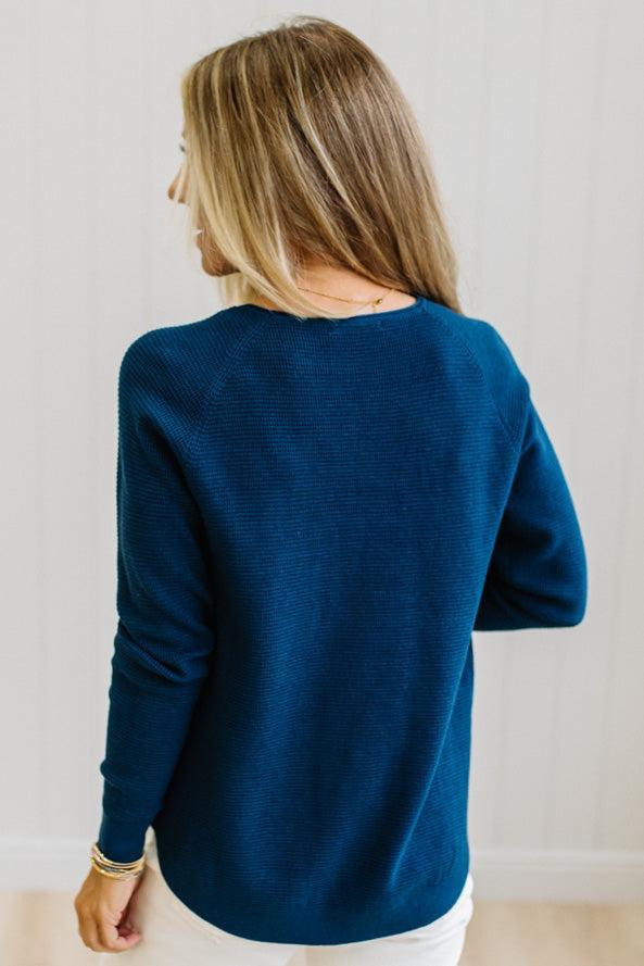 A woman with blonde hair is shown from behind wearing a textured, ribbed knit long-sleeve sweater in a deep teal blue color, paired with white pants.