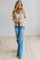 A woman modeling a beige and white striped cardigan over a white tank top and blue jeans, standing in a bright room with light wood floors and white paneled walls.