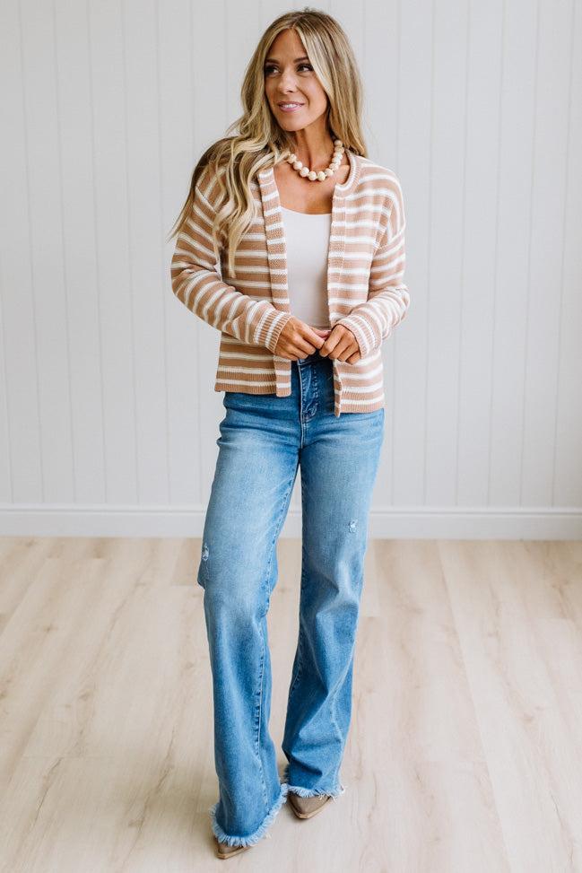 A woman modeling a beige and white striped cardigan over a white tank top and blue jeans, standing in a bright room with light wood floors and white paneled walls.
