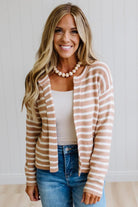 A smiling woman with blonde hair models a beige and white horizontally striped knit cardigan over a white tank top and blue jeans, standing against a white paneled wall.