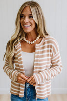 A woman with blonde hair wearing a beige and white striped knit cardigan over a white tank top and blue jeans, accessorized with a chunky white beaded necklace.