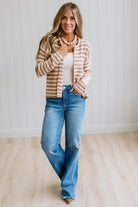 A woman modeling a beige and white striped cardigan over a white tank top and blue jeans, standing in a bright room with light wood floors and white paneled walls.