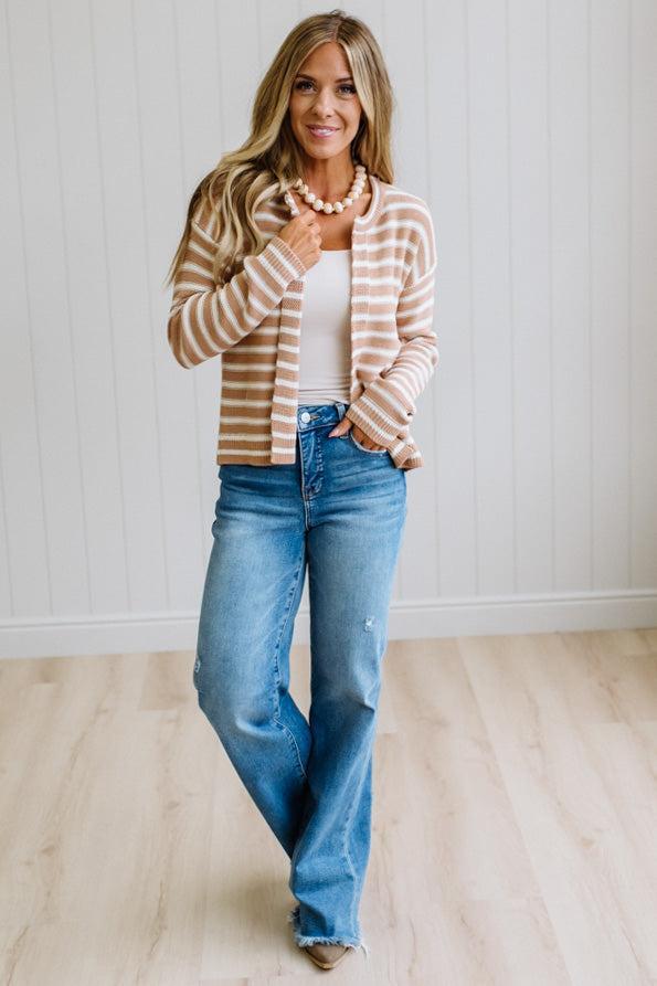 A woman modeling a beige and white striped cardigan over a white tank top and blue jeans, standing in a bright room with light wood floors and white paneled walls.