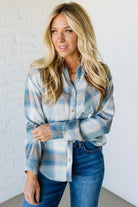 Woman wearing a blue plaid shirt and jeans against a white brick wall.