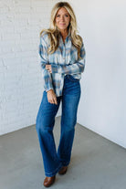 Woman wearing a blue plaid shirt and jeans standing against a white brick wall.