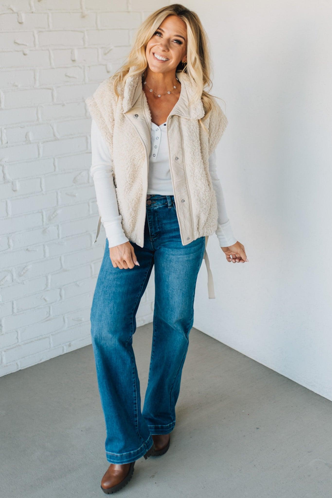 Woman wearing a beige vest, white shirt, blue jeans, and brown shoes against a white brick wall.
