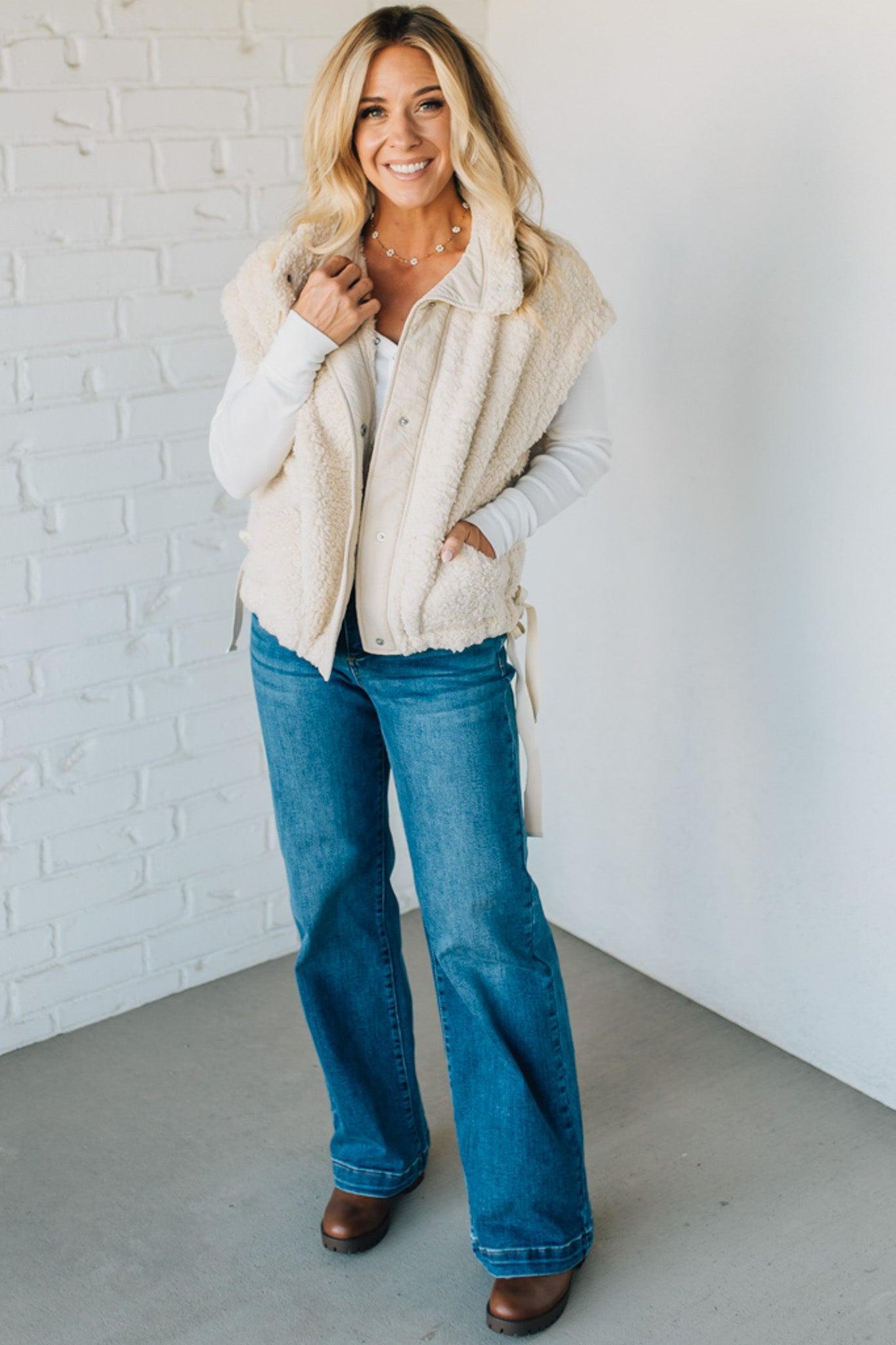 Woman wearing a cream vest, white sweater, blue jeans, and brown shoes against a white brick wall.
