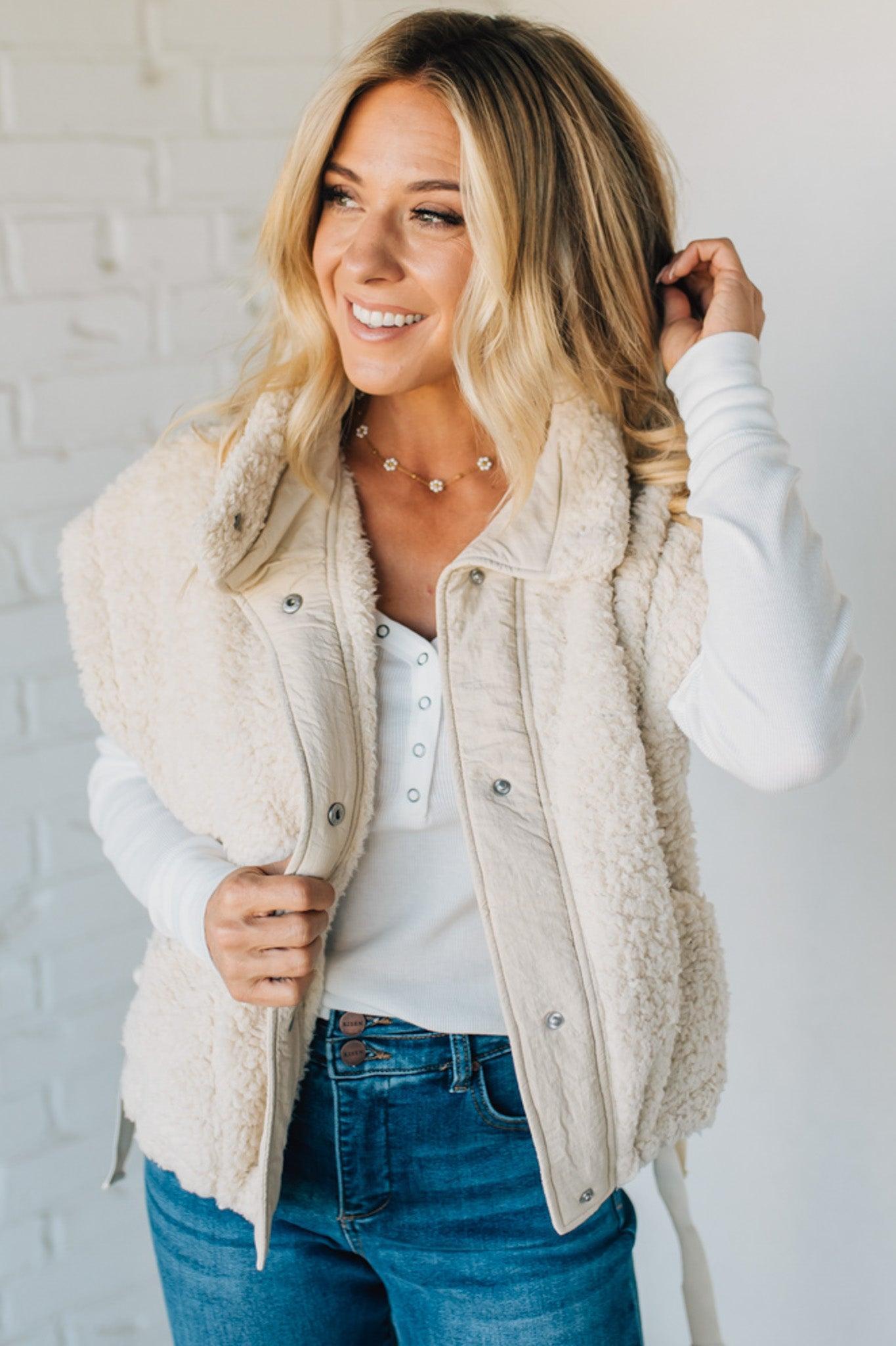 Woman wearing a cream-colored vest over a white shirt and blue jeans against a white brick wall.