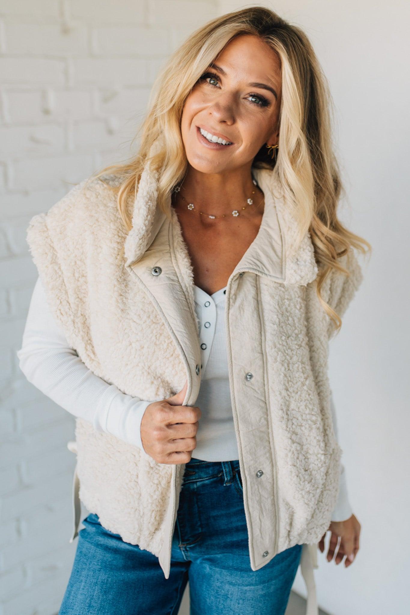 Woman wearing a beige vest over a white shirt and blue jeans against a white brick wall.