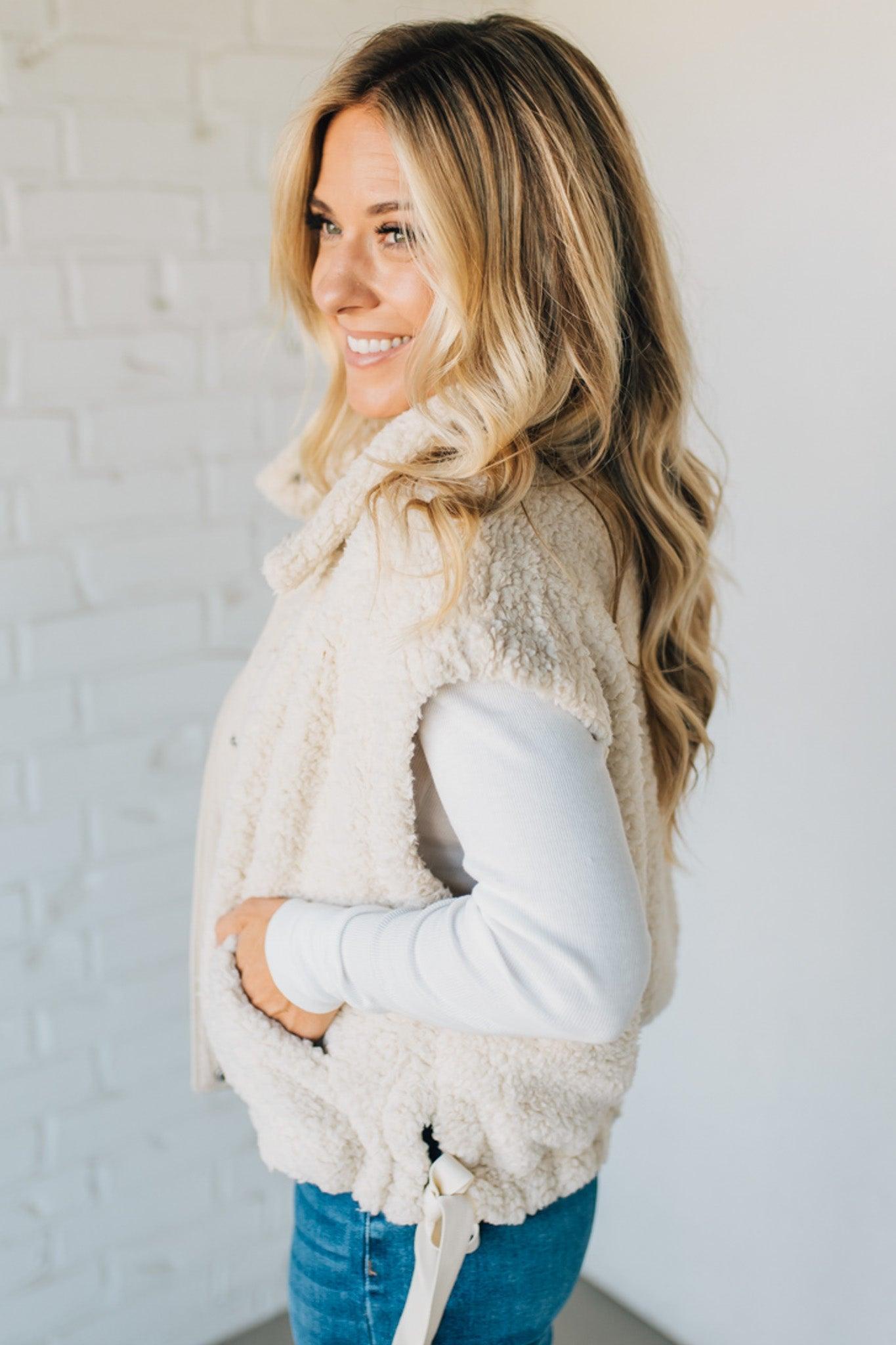 Woman wearing a beige sherpa vest against a white brick wall
