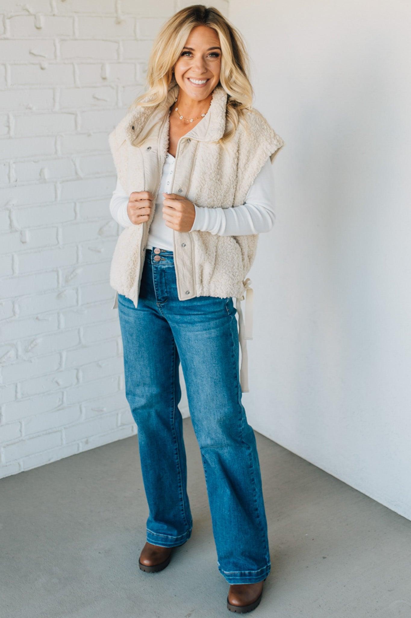 Woman wearing a beige vest, white shirt, blue jeans, and brown shoes against a white brick wall.