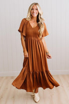 A woman modeling a rust-brown tiered maxi dress with flutter sleeves and a smocked bodice, standing in front of a white paneled wall and light wood floor.