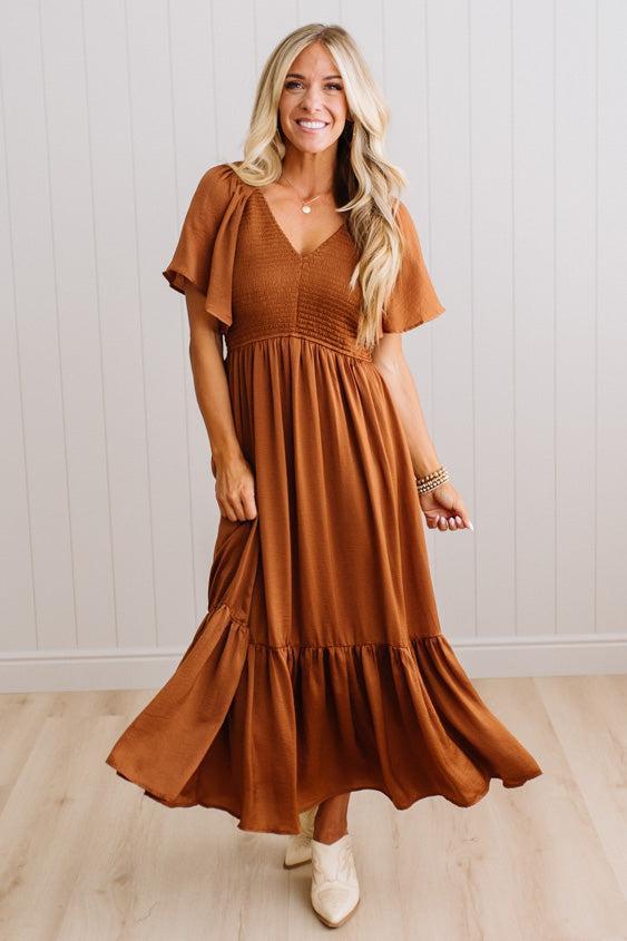 A woman modeling a rust-brown tiered maxi dress with flutter sleeves and a smocked bodice, standing in front of a white paneled wall and light wood floor.