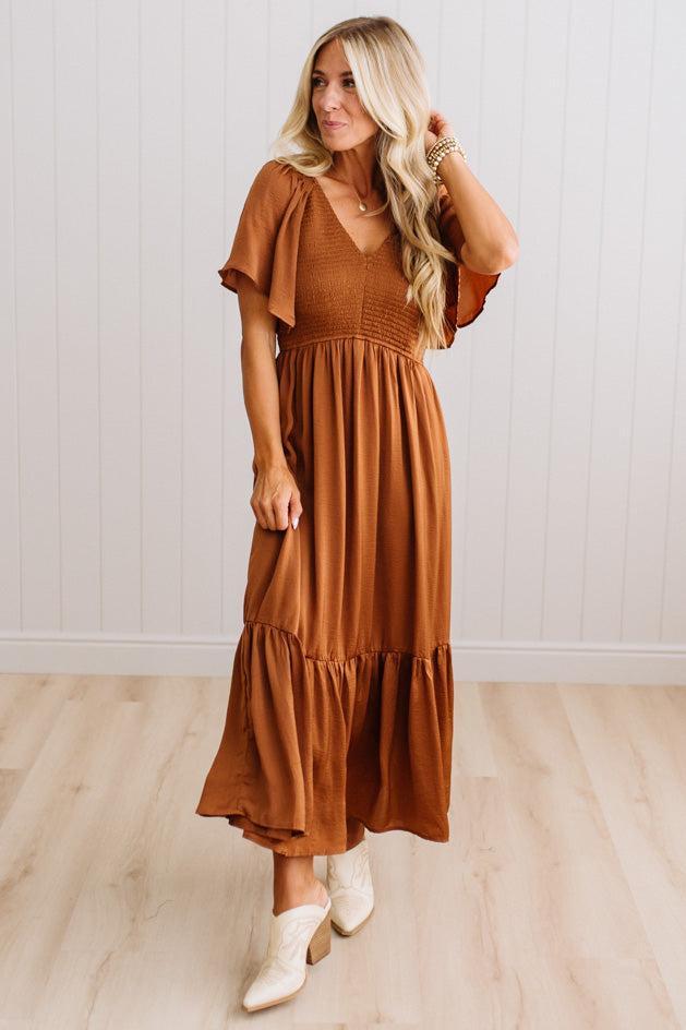 A woman modeling a rust-brown tiered maxi dress with flutter sleeves and a smocked bodice, paired with white heeled boots, standing in a room with light wood floors and white paneled walls.