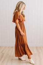 A woman modeling a rust-brown tiered maxi dress with flutter sleeves and cream ankle boots, standing in a room with light wood floors and white paneled walls.