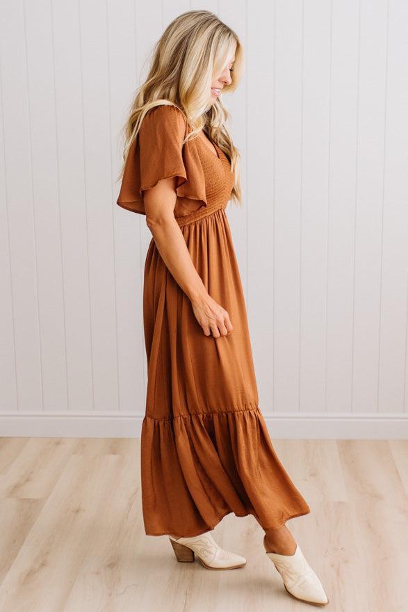 A woman modeling a rust-brown tiered maxi dress with flutter sleeves and cream ankle boots, standing in a room with light wood floors and white paneled walls.