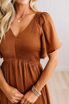 A close-up of a woman wearing a rust-brown smocked dress with flutter sleeves and a V-neckline, paired with layered gold bracelets and a delicate gold necklace.