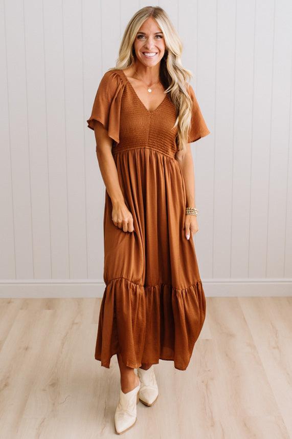 A woman modeling a rust-brown tiered midi dress with flutter sleeves and a smocked bodice, paired with cream ankle boots, standing in front of a white paneled wall and light wood floor.