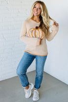 Woman wearing a beige sweater with a pumpkin design against a white brick wall.