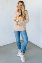 Woman wearing a beige sweater with a pumpkin design, holding a small pumpkin, against a white brick wall.