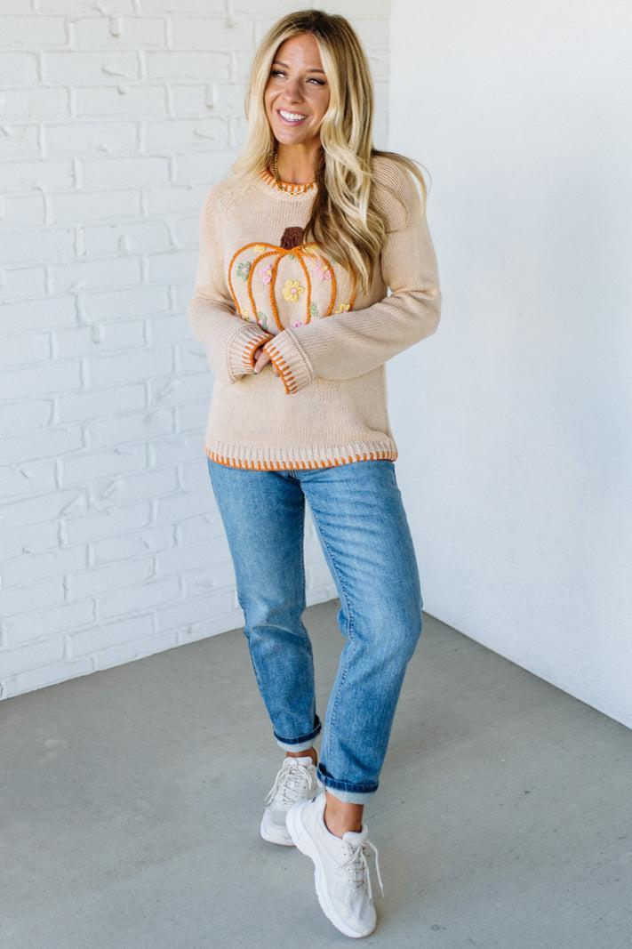 Woman wearing a beige sweater with a pumpkin design, holding a small pumpkin, against a white brick wall.