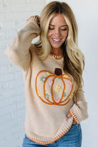 Woman wearing a beige sweater with a pumpkin design against a white brick wall.