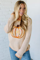 Woman wearing a beige sweater with a pumpkin design against a white brick wall.