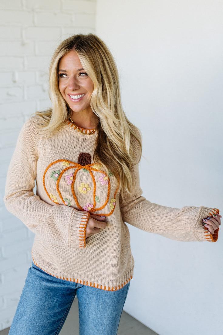 Woman wearing a beige sweater with a pumpkin design, holding a decorative pumpkin.