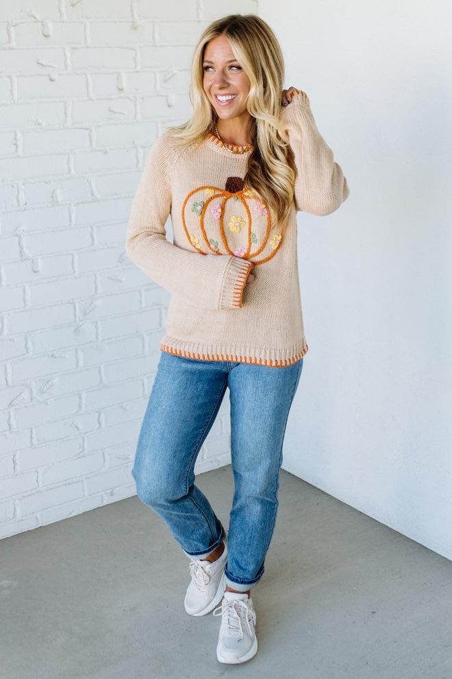 Woman wearing a beige sweater with a pumpkin design against a white brick wall.