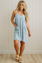 Woman wearing a light blue dress with pockets against a white paneled wall.