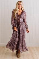 A woman modeling a long-sleeve, V-neck maxi dress with a brown and purple floral pattern, standing in front of a white paneled wall and light wood floor.