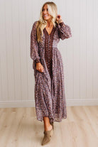 A woman modeling a long-sleeve, V-neck maxi dress with a dense floral pattern in muted purple, brown, and beige tones, paired with taupe ankle boots, standing against a white paneled wall and light wood floor.