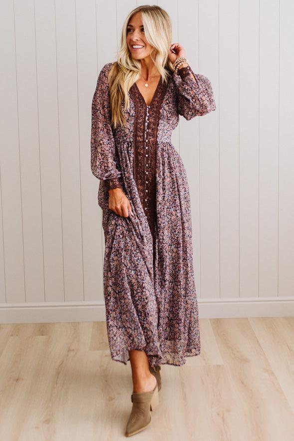 A woman modeling a long-sleeve, V-neck maxi dress with a dense floral pattern in muted purple, brown, and beige tones, paired with taupe ankle boots, standing against a white paneled wall and light wood floor.
