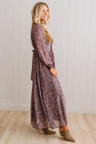 A woman modeling a long-sleeve maxi dress with a dense floral and abstract print in muted purple, pink, and brown tones, paired with brown ankle boots, standing against a white paneled wall and light wood floor.