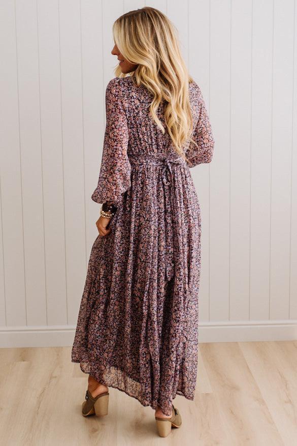 A woman wearing a long-sleeved, knee-length floral maxi dress in a dusty rose and purple floral print, shown from behind against a white paneled wall and light wood floor.