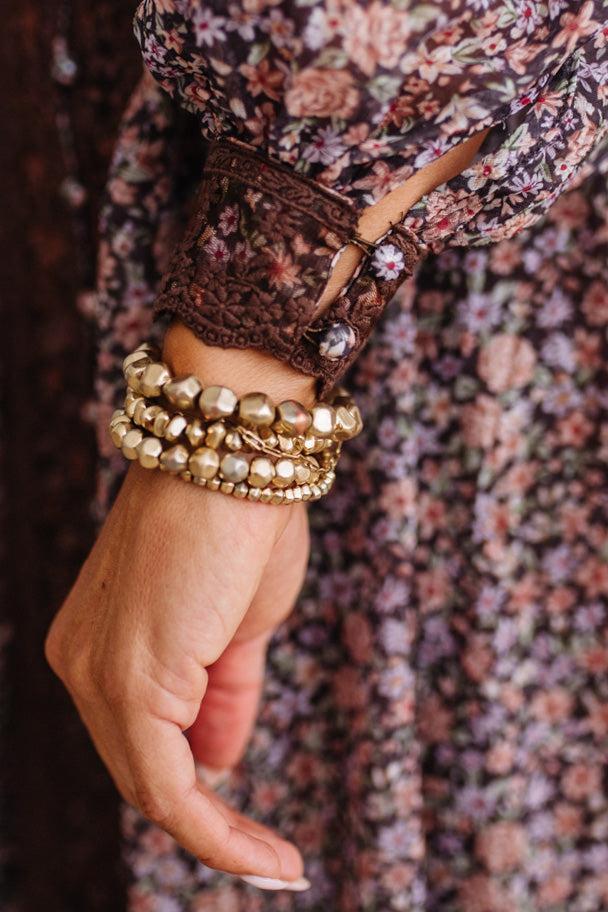A close-up of a person's hand and wrist wearing multiple stacked gold-toned beaded bracelets, with a floral dress and dark lace cuff visible.