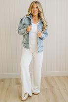 Woman wearing a denim jacket over a white top and light-colored pants indoors.