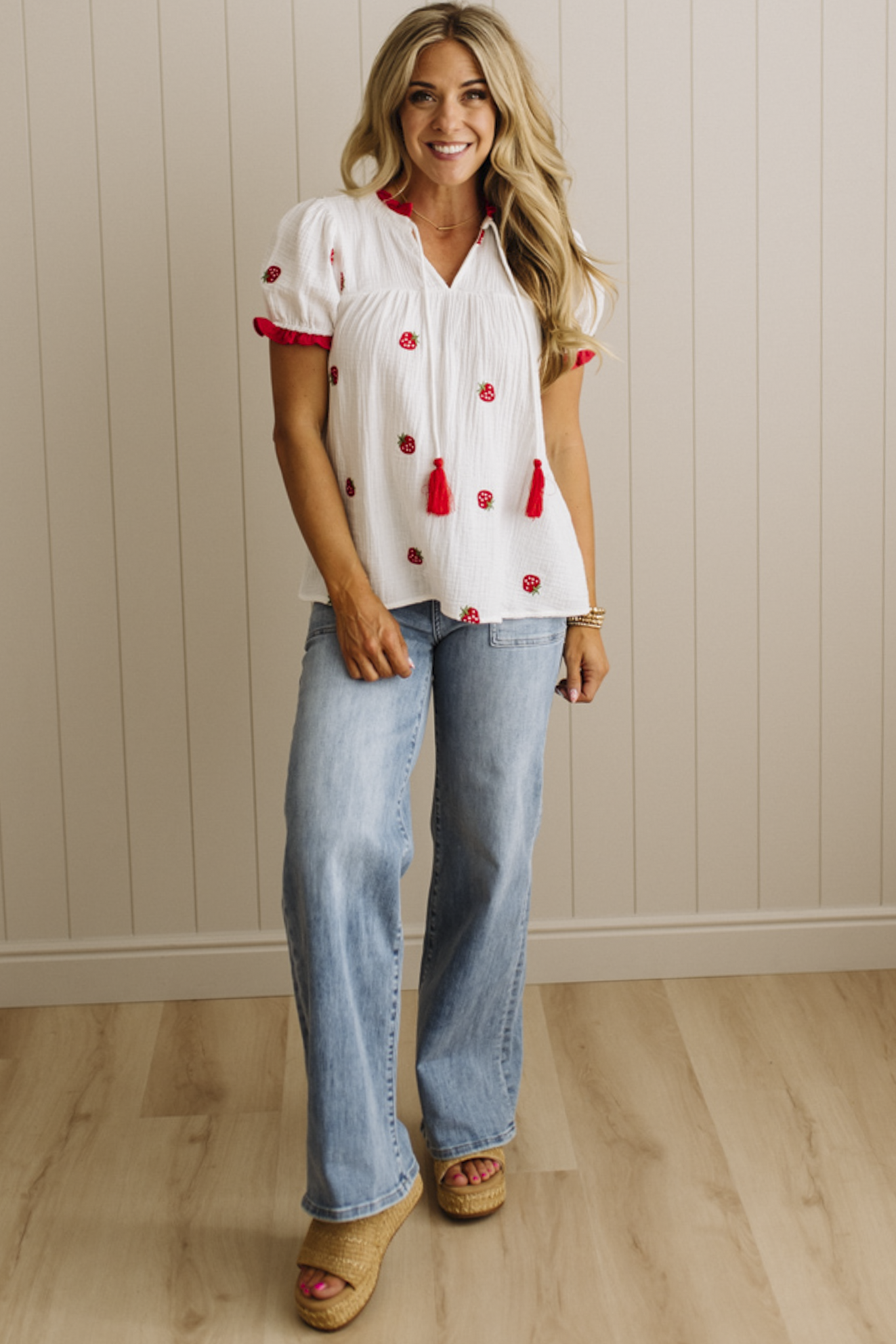Woman wearing a white blouse with red accents and blue jeans against a beige wall.