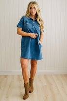Woman wearing a denim dress and brown boots against a white paneled wall.
