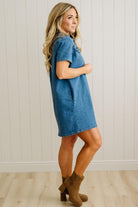 Woman wearing a blue denim dress with brown boots against a white paneled wall.