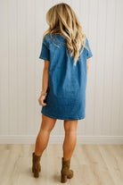 Woman wearing a blue denim dress with brown boots against a white paneled wall.