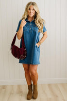 Woman wearing a blue dress holding a maroon handbag against a white paneled wall.